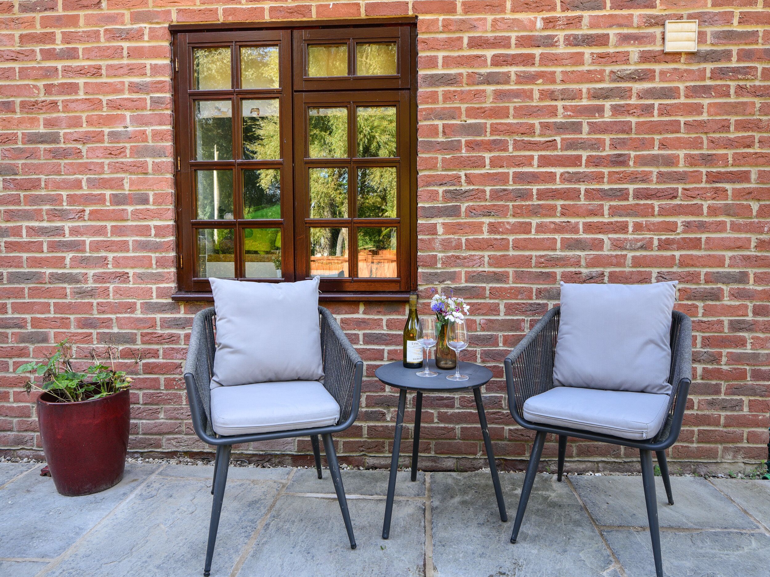 Two wicker chairs with cushions, a small round table, and a potted plant against a brick wall with a window.