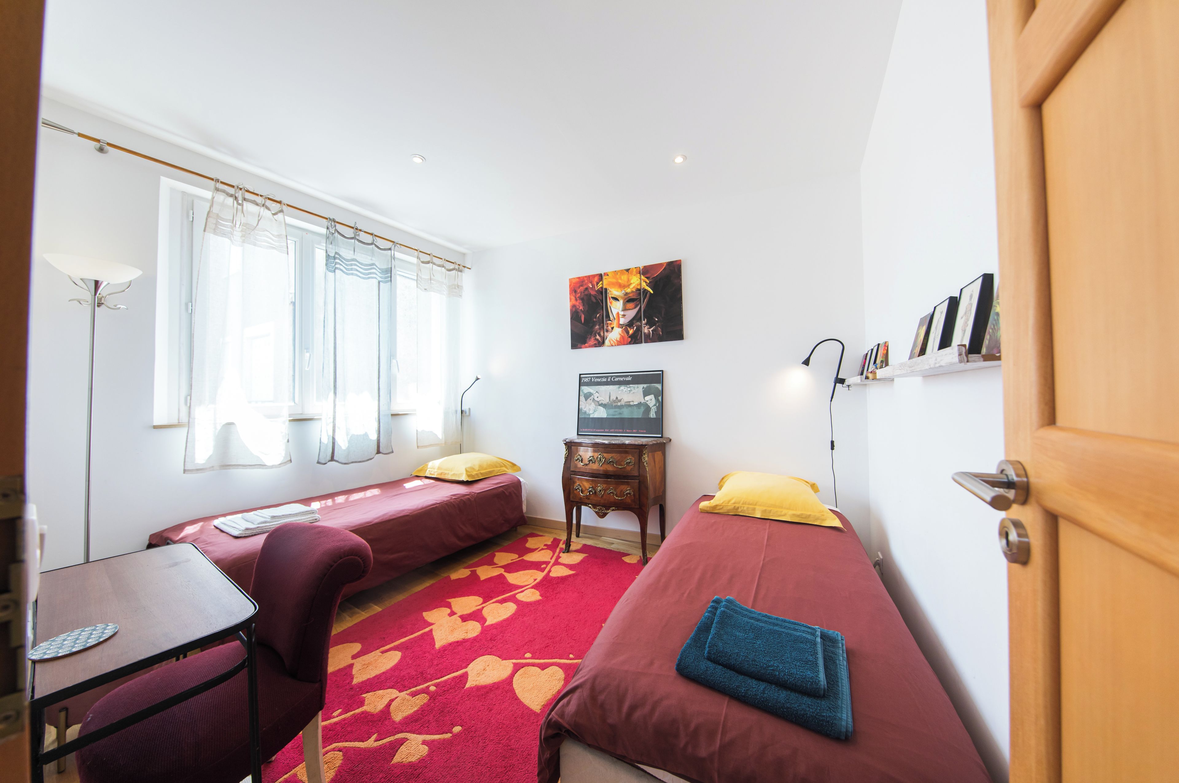 A room with two single beds, a small table, a chair, a lamp, a picture frame, and a door.