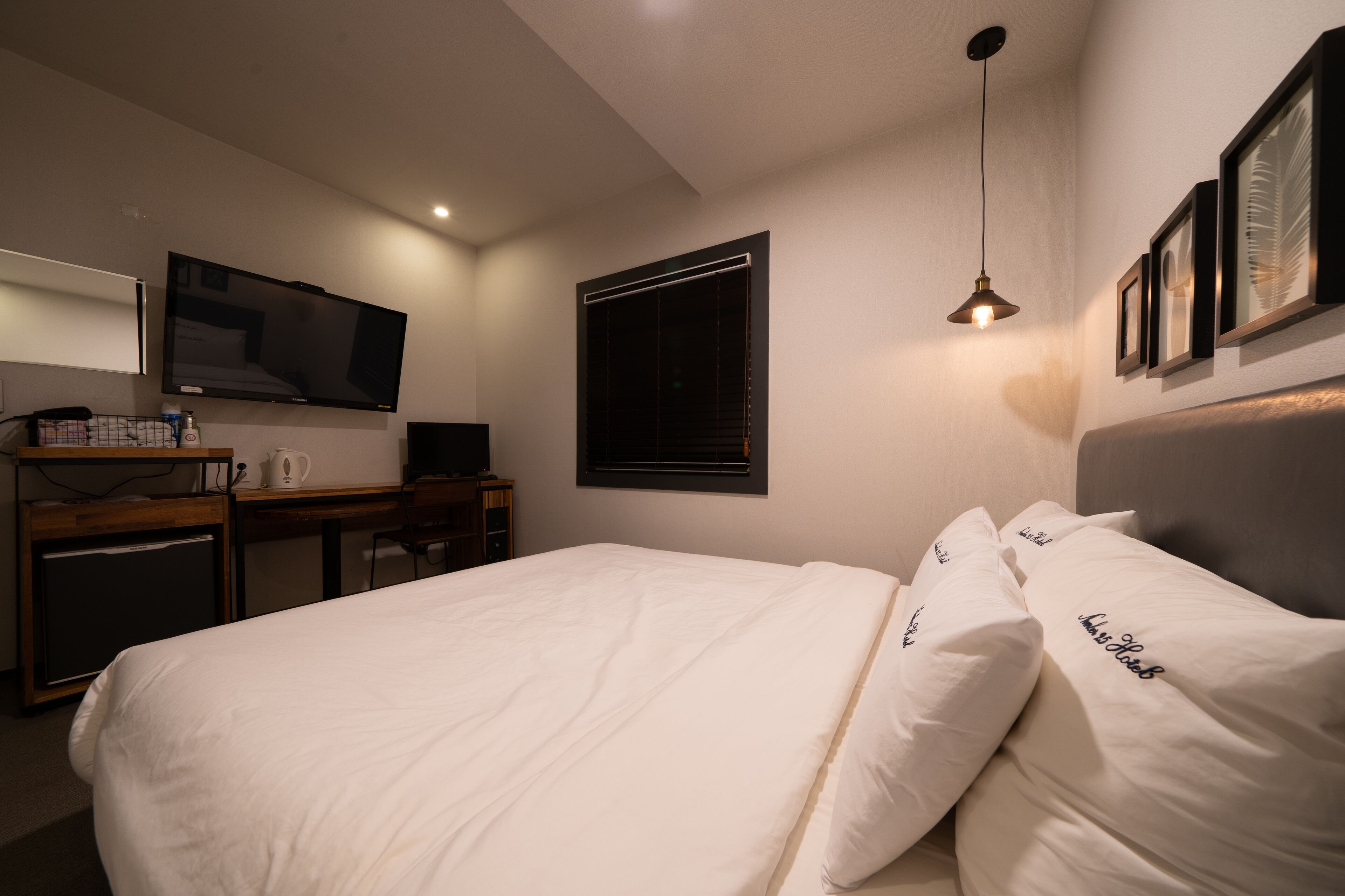 Ein Hotelzimmer mit einem Bett, einem Schreibtisch mit Computer, einem Fernseher und einem Fenster mit Jalousien.