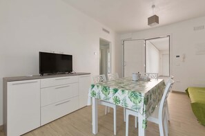 Quarto moderno e bem iluminado com uma mesa de jantar, cadeiras brancas, uma TV de tela plana em um móvel e um espelho grande na parede.
