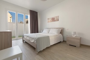 Quarto com cama, mesa de cabeceira, escrivaninha e porta de vidro deslizante para área externa.