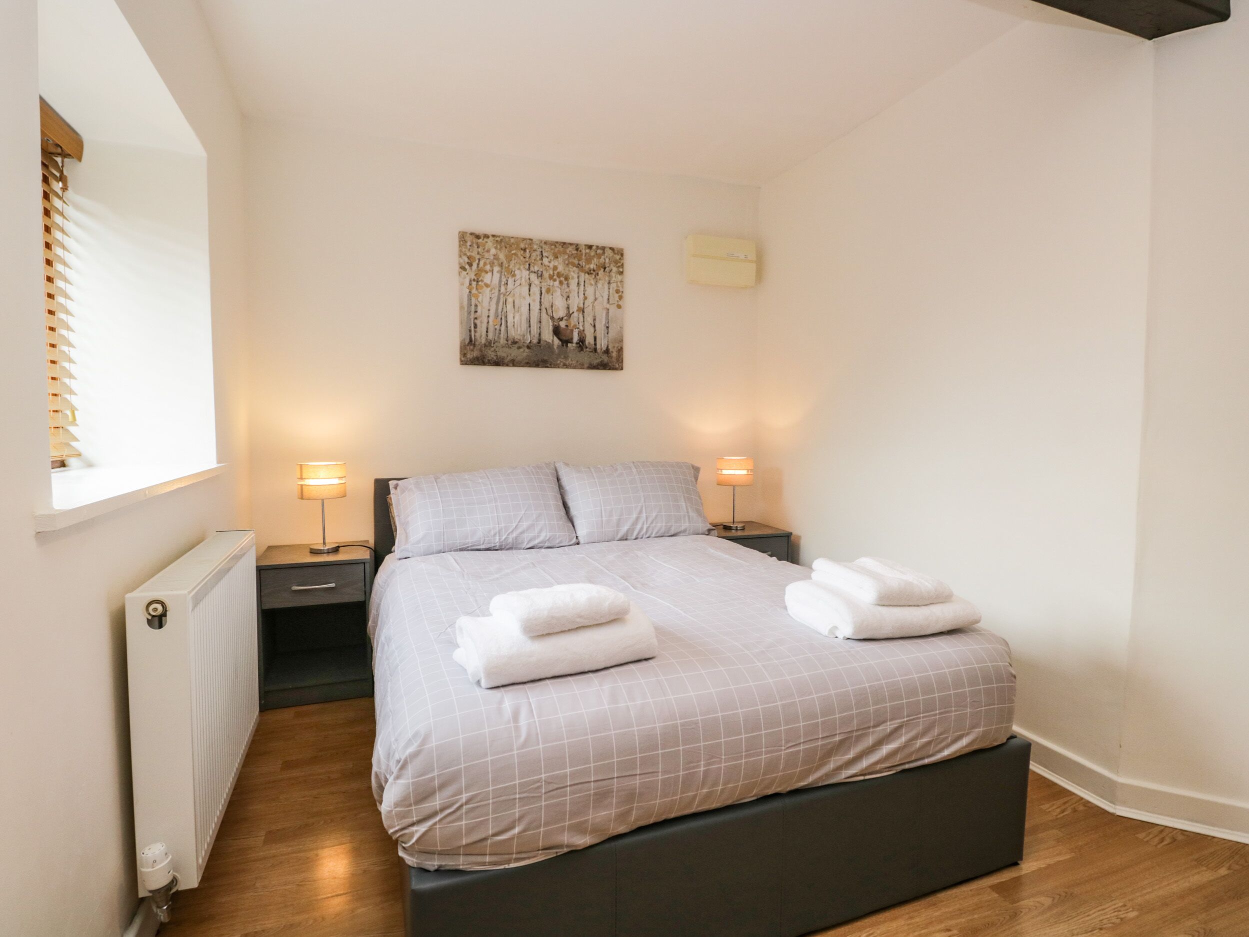 A neatly made bed with a checkered bedspread, two bedside lamps, a radiator, and a window with blinds.
