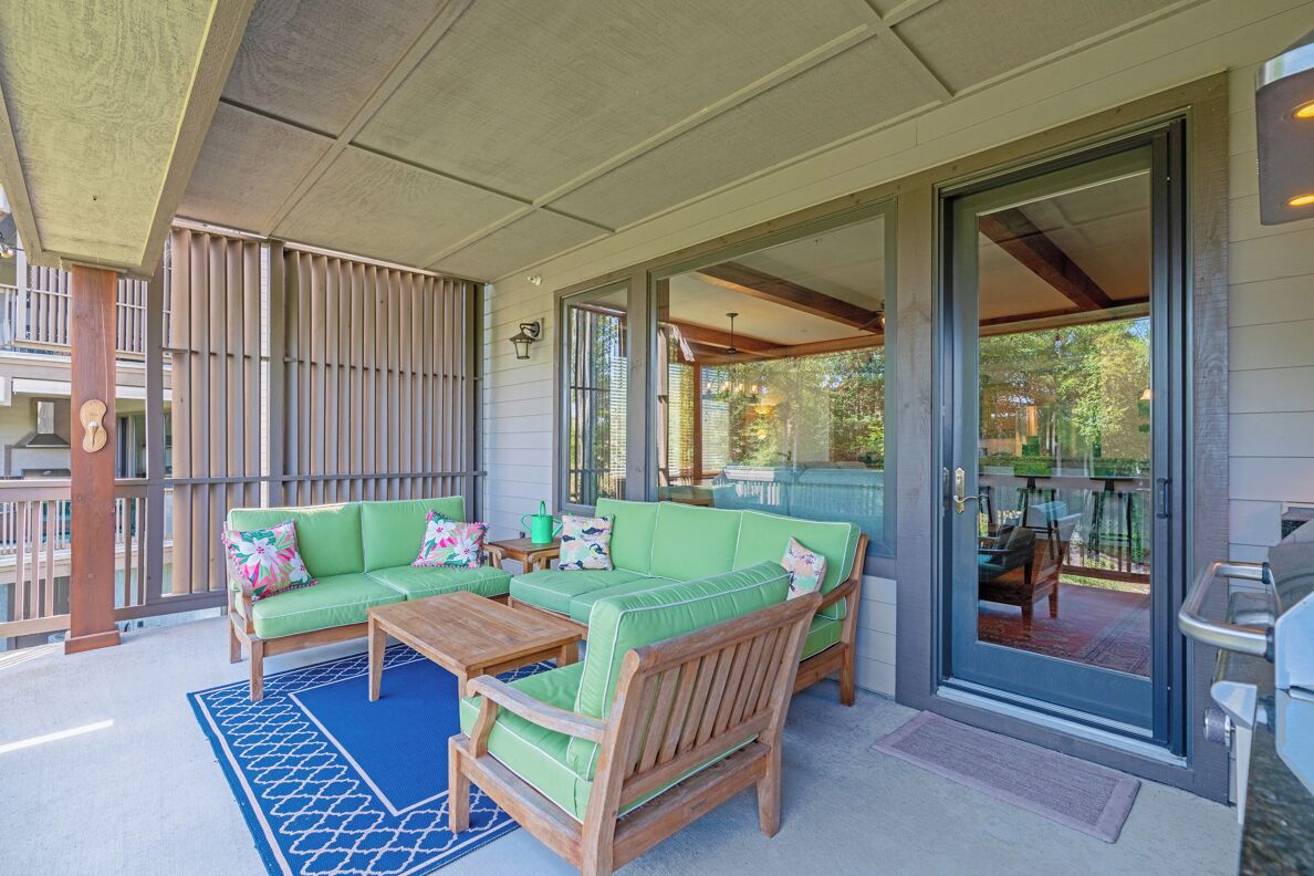 A covered outdoor seating area with a green sofa and wooden coffee table, a grill, and a view of the backyard.