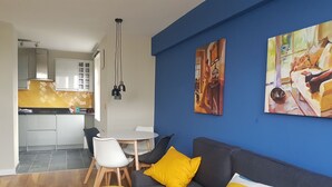 Un salon moderne avec un canapé gris, des coussins jaunes et un plaid coloré. On y trouve également un coin repas avec une table et des chaises blanches, et une cuisine en arrière-plan.