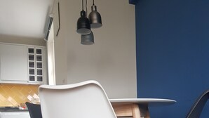 Un coin repas avec une table blanche et quatre chaises, dont deux blanches et deux noires, avec des pieds en bois. Des lampes suspendues éclairent l’espace.