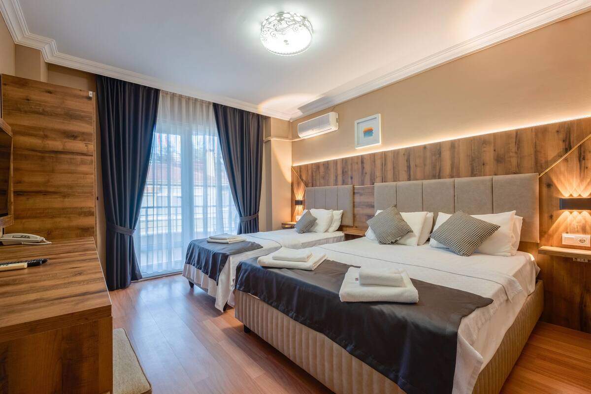 A hotel room with two beds, a wooden desk, and a large window with curtains.