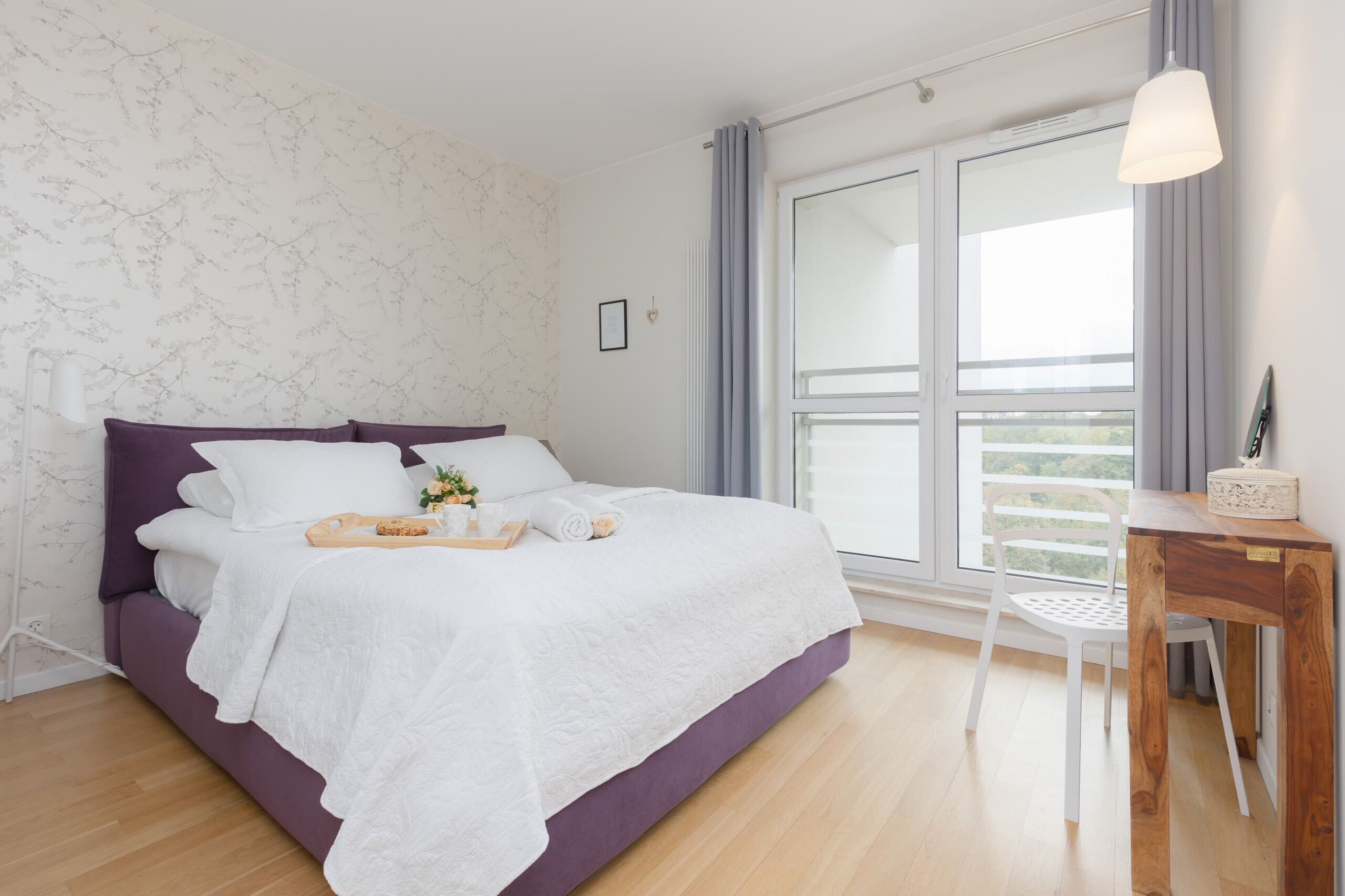 A modern bedroom with a large bed, a wooden desk, and a view of the outdoors.