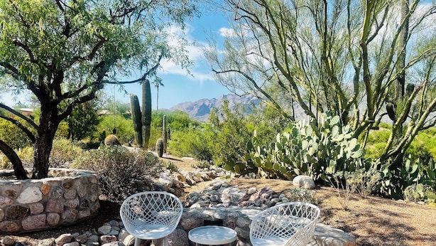 An outdoor seating area with two white metal chairs and a small round table, surrounded by rocks and cacti, with a view of mountains in the background.