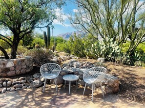 An outdoor seating area with two white metal chairs and a small round table, surrounded by rocks and cacti, with a view of mountains in the background. - Quiet desert getaway with beautiful views and heated pool, pet-friendly (Tucson)