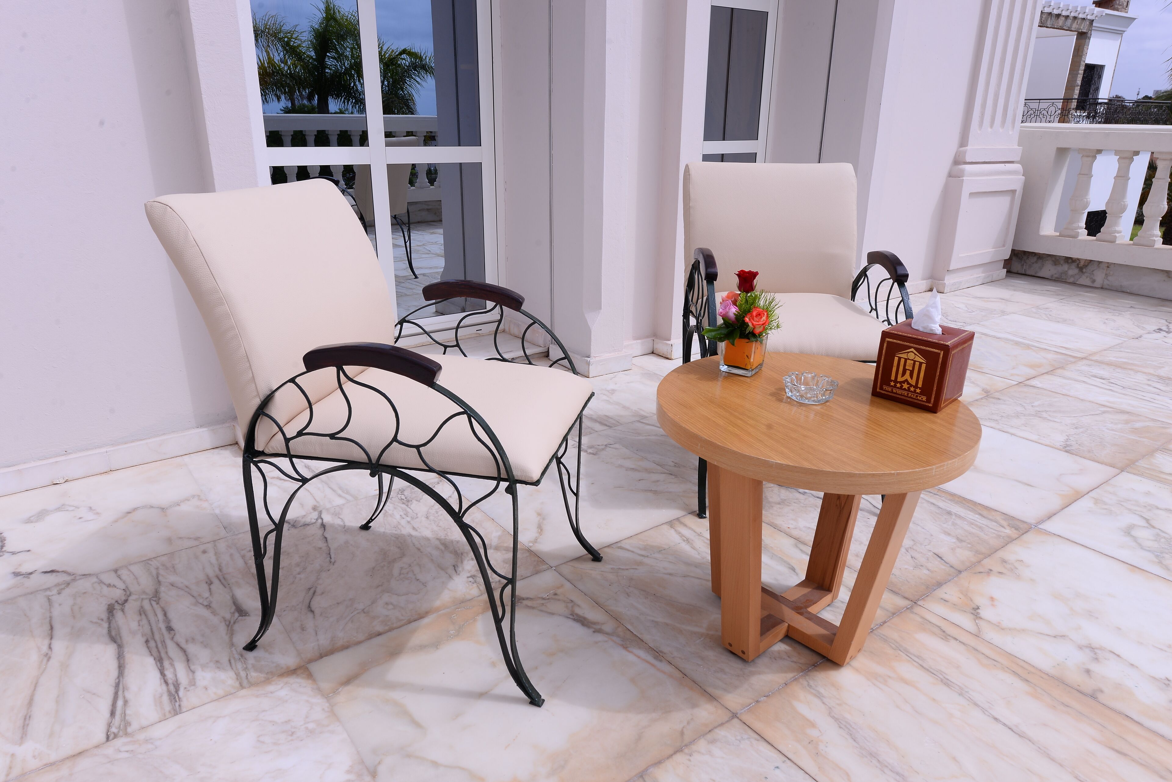 Een terras met twee stoelen, een ronde houten tafel en een vaas met bloemen.