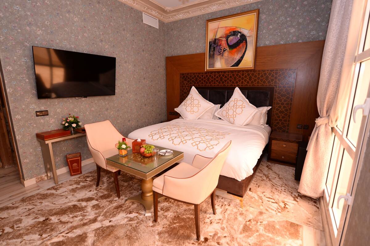A hotel room with a bed, a TV, a table with chairs, and a small desk with a vase of flowers.