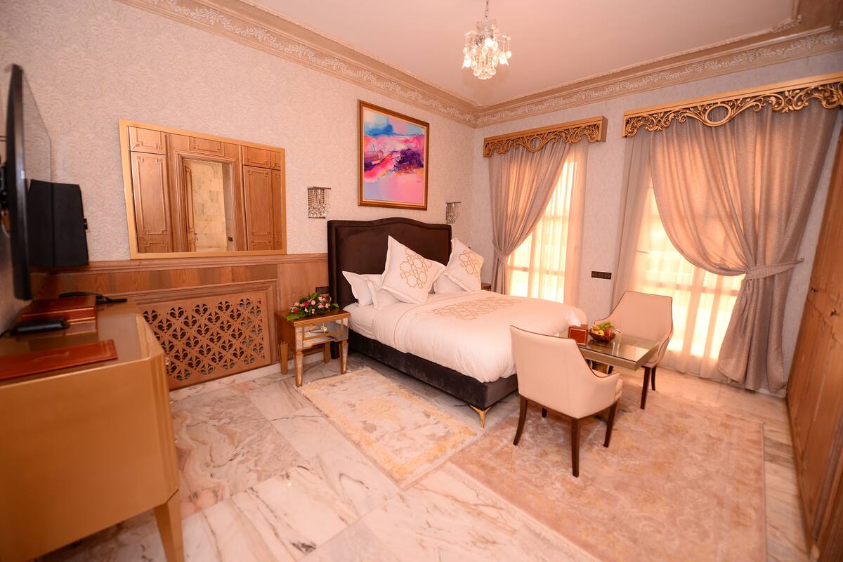 A well-appointed bedroom with a large bed, a desk, and a chair, featuring a chandelier and a large window with curtains.