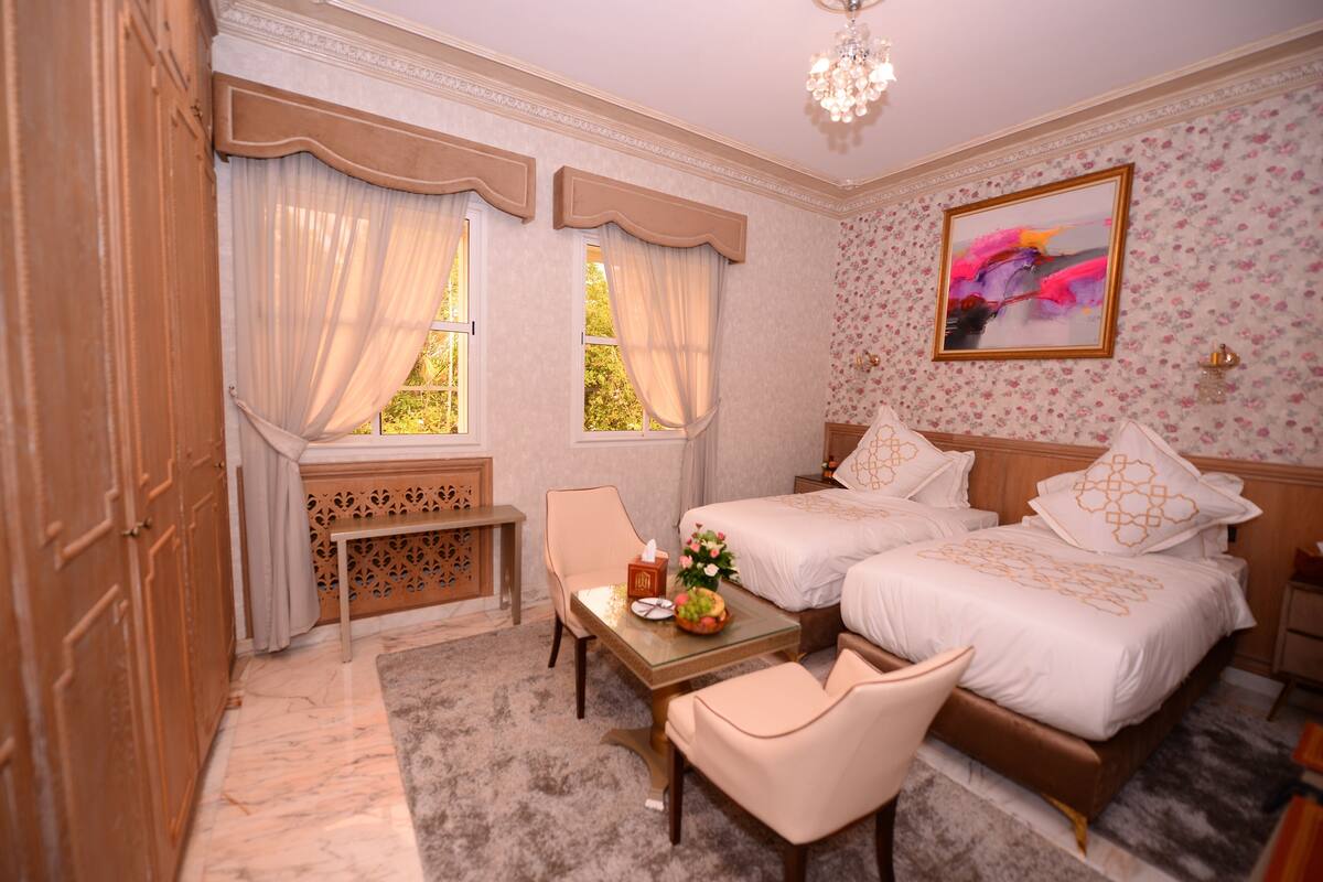 A hotel room with two beds, a small table with a vase of flowers, a chair, and a decorative wall piece.