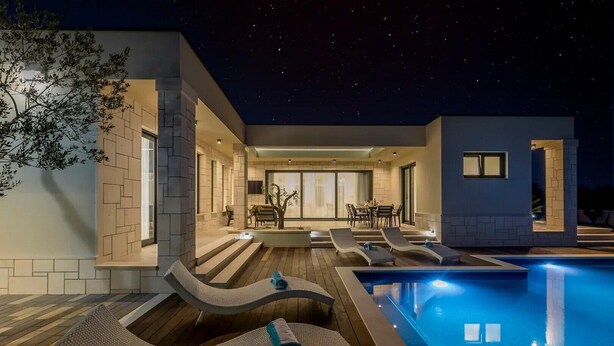 Un área de piscina moderna al aire libre con sillones de descanso y comedor bajo un cielo nocturno.
