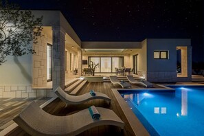Et moderne poolområde udendørs med liggestole og et spiseområde under en nattehimmel.