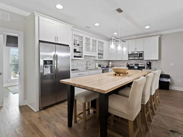 Una cocina moderna con gabinetes blancos, electrodomésticos de acero inoxidable, una mesa de comedor de madera y luces colgantes.