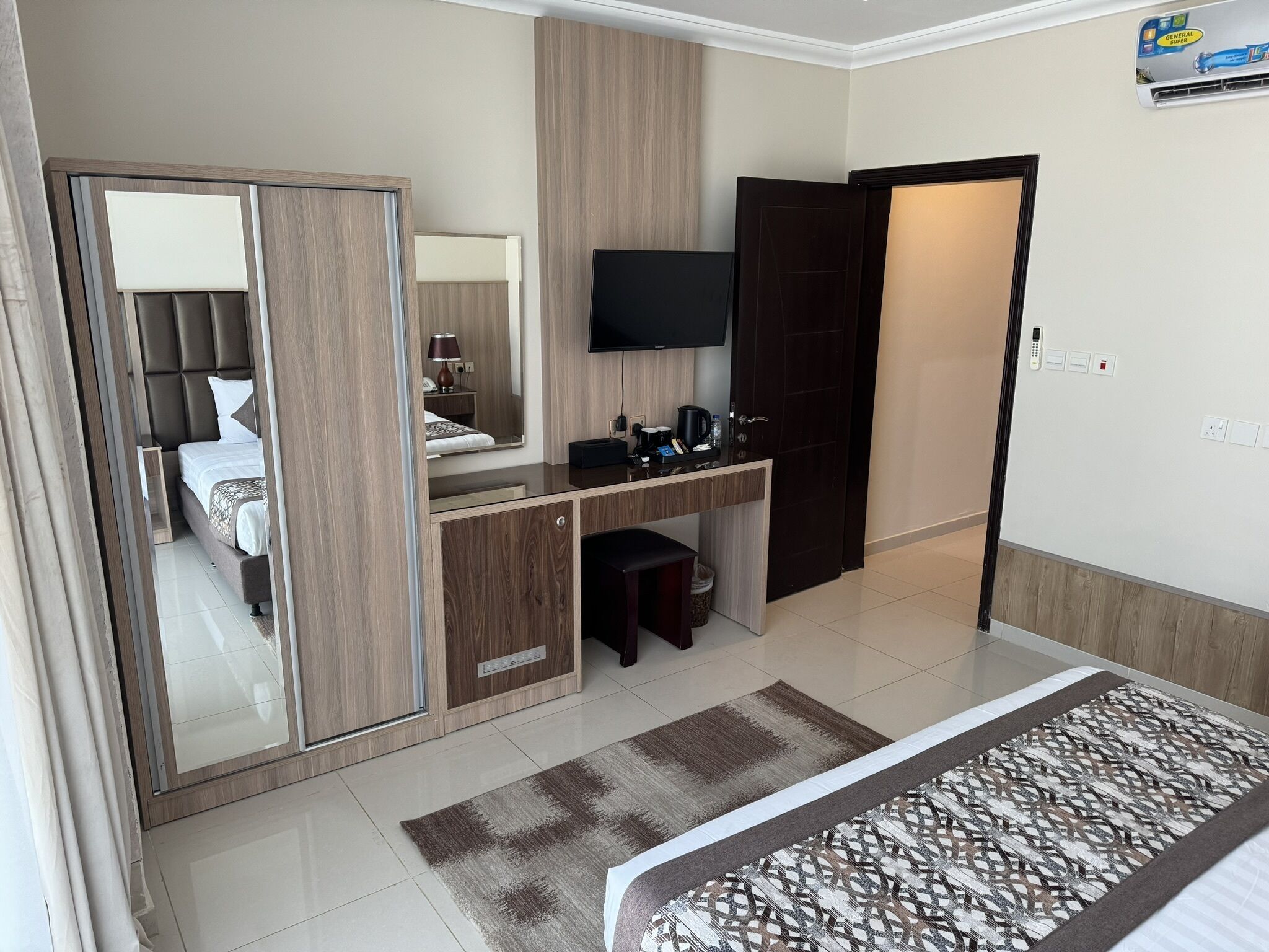 A hotel room with a bed, a desk with a flat-screen TV, a mirror, a wardrobe, and a bathroom door.