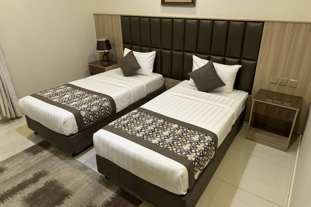 Quarto duplo com duas camas, mesa de cabeceira, abajur e pequena mesa com abajur.