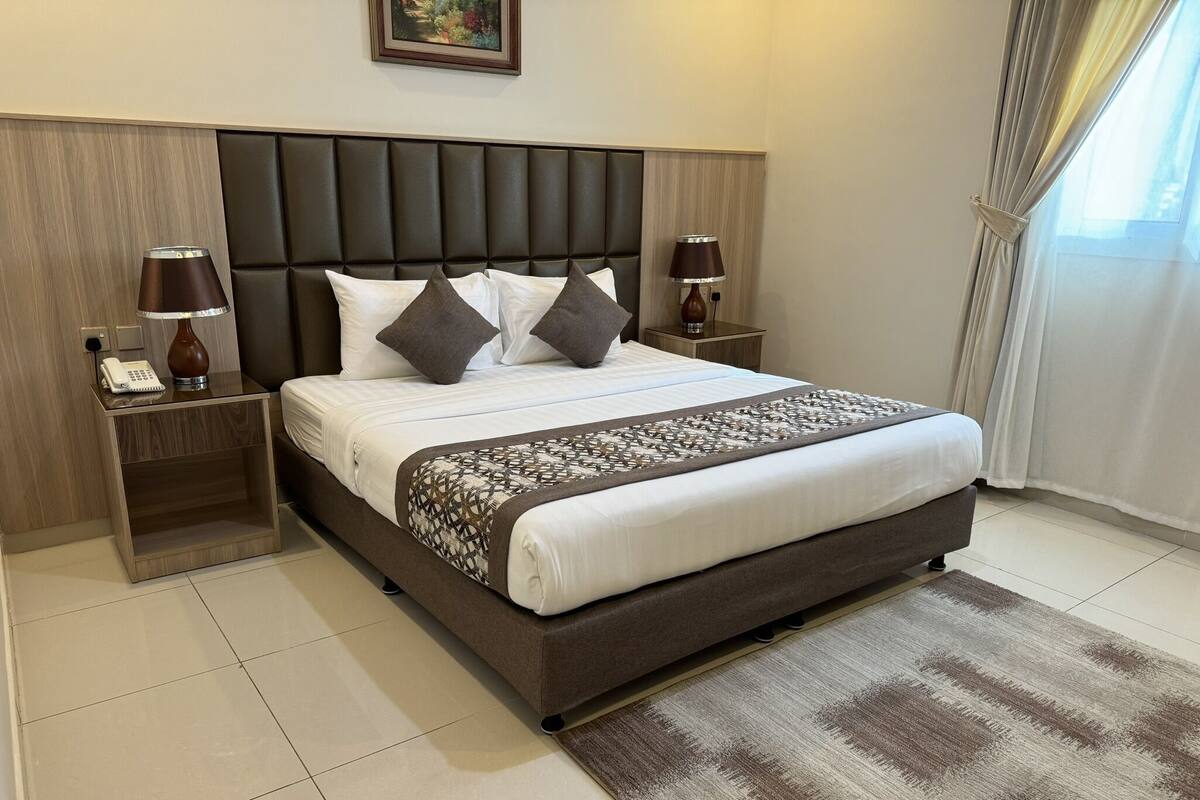 Quarto de hotel moderno com uma cama grande, duas mesas de cabeceira com luminárias, cabeceira de madeira e manta decorativa.