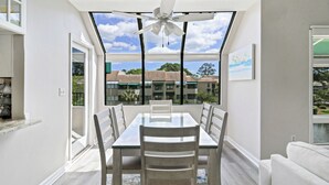 A dining area with a table and chairs, a ceiling fan, and large windows offering a view of buildings and trees. - 2 bedroom villa just steps to the pool with a fantastic lagoon view! (Hilton Head Island)