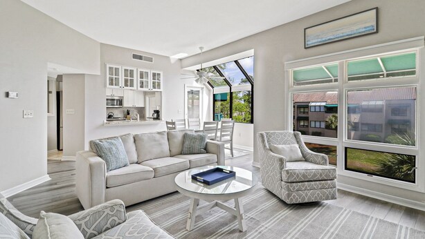 A modern living room with a sofa, armchairs, and a coffee table. There is a kitchen area with white cabinets and a dining area with a table and chairs. Large windows provide views of the outside. - 2 bedroom villa just steps to the pool with a fantastic lagoon view! (Hilton Head Island)
