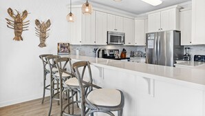 A modern kitchen with white cabinets, a stainless steel refrigerator, a microwave, and a bar area with wooden chairs. - CottageVacations4u "The Great Escape"-ocean view villa-pool club-direct beach access (Ocean Isle Beach)