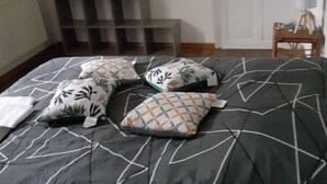 Makuuhuone, jossa on sänky, tyynyjä, patteri, valkoinen ovi ja pieni pöytä, jossa on lamppu.