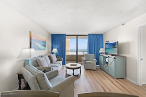 A living room with a sofa set, a coffee table, a TV, and a view of the ocean through large windows.