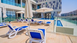 A rooftop pool area with sun loungers and umbrellas, a modern building with large windows, and a clear view of the city skyline. - Nasma Luxury Stays - Al Raha Lofts 2 (Abu Dhabi)
