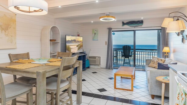A dining area with a wooden table and chairs, a sofa, and a balcony with a view of the sea. - Beachers 421: stunning oceanfront, steps to pool, pet friendly. (St. Augustine)