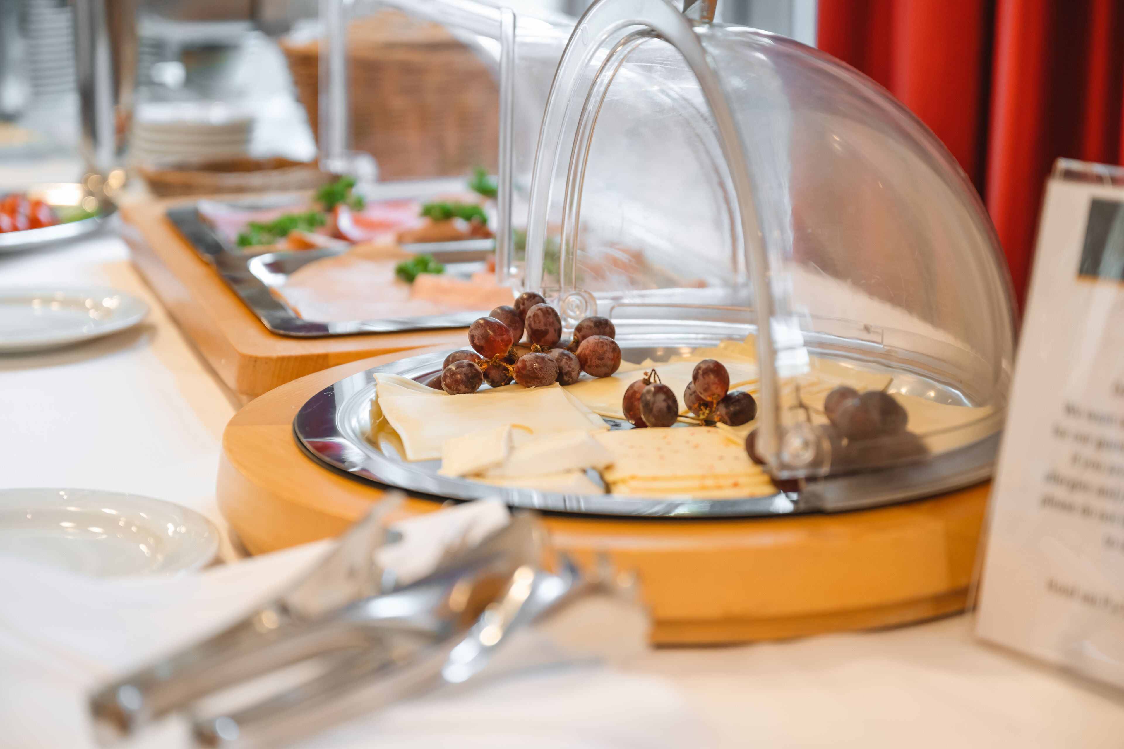 Montaje tipo bufé con diversos alimentos bajo una cubierta de vidrio, incluyendo queso, galletas y uvas.