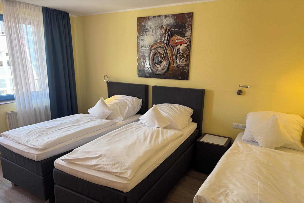 A hotel room with two single beds, a nightstand, a wall-mounted painting, and a window with curtains.