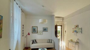 Ein geräumiges Wohnzimmer mit einem weißen Sofa, einem kleinen Tisch und einem weißen Sessel. An der Wand befinden sich gerahmte Bilder und eine Stehlampe.