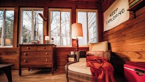 L’intérieur chaleureux d’un chalet avec une commode en bois, un fauteuil en osier, une valise rouge et une pancarte sur laquelle on peut lire : « CONTINUEZ D’EXPLORER ».