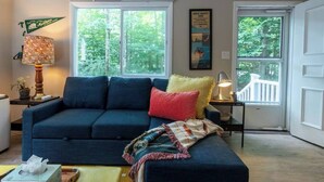 Un salon avec un canapé bleu foncé, une petite table avec une lampe et une fenêtre donnant sur des arbres.