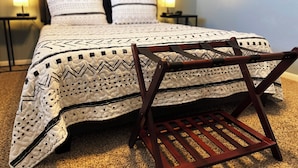 Cama bem arrumada com colcha estampada e almofadas, uma cadeira de madeira e duas luminárias de cabeceira.