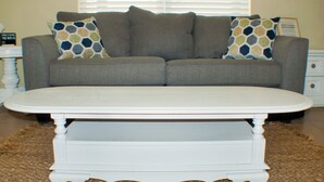 Un salon avec un canapé gris, deux coussins décoratifs à motifs, une table basse blanche et un tapis marron.