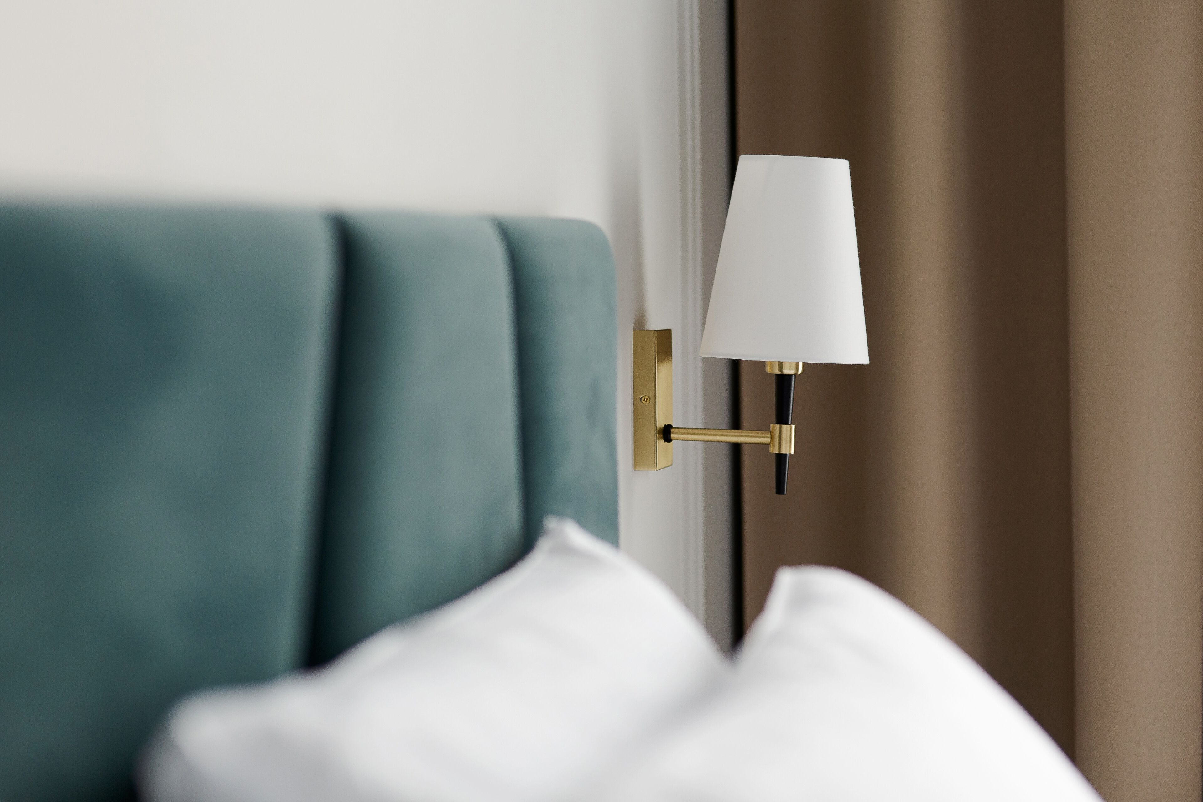 A modern hotel room with a teal upholstered headboard, a gold-framed wall-mounted lamp, and white bedding.