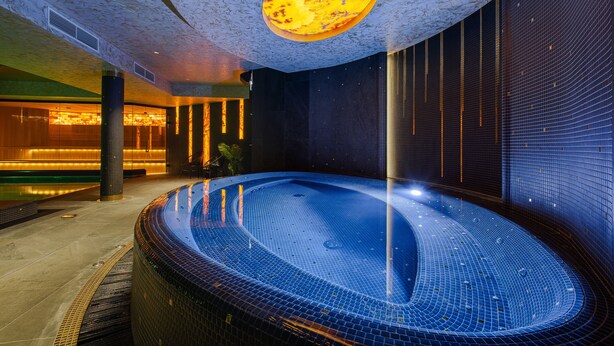 A modern indoor swimming pool with a unique ceiling design, surrounded by dark tiled walls and a warm ambient light. - VICTORIA Hotel Klaipėda (Klaipeda)