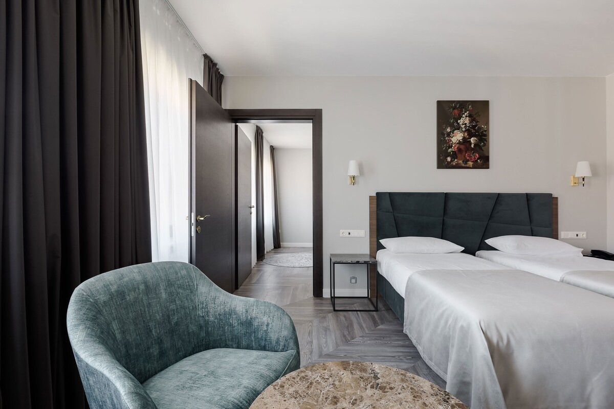 A hotel room with a large bed, a chair, a small table, and a painting on the wall.