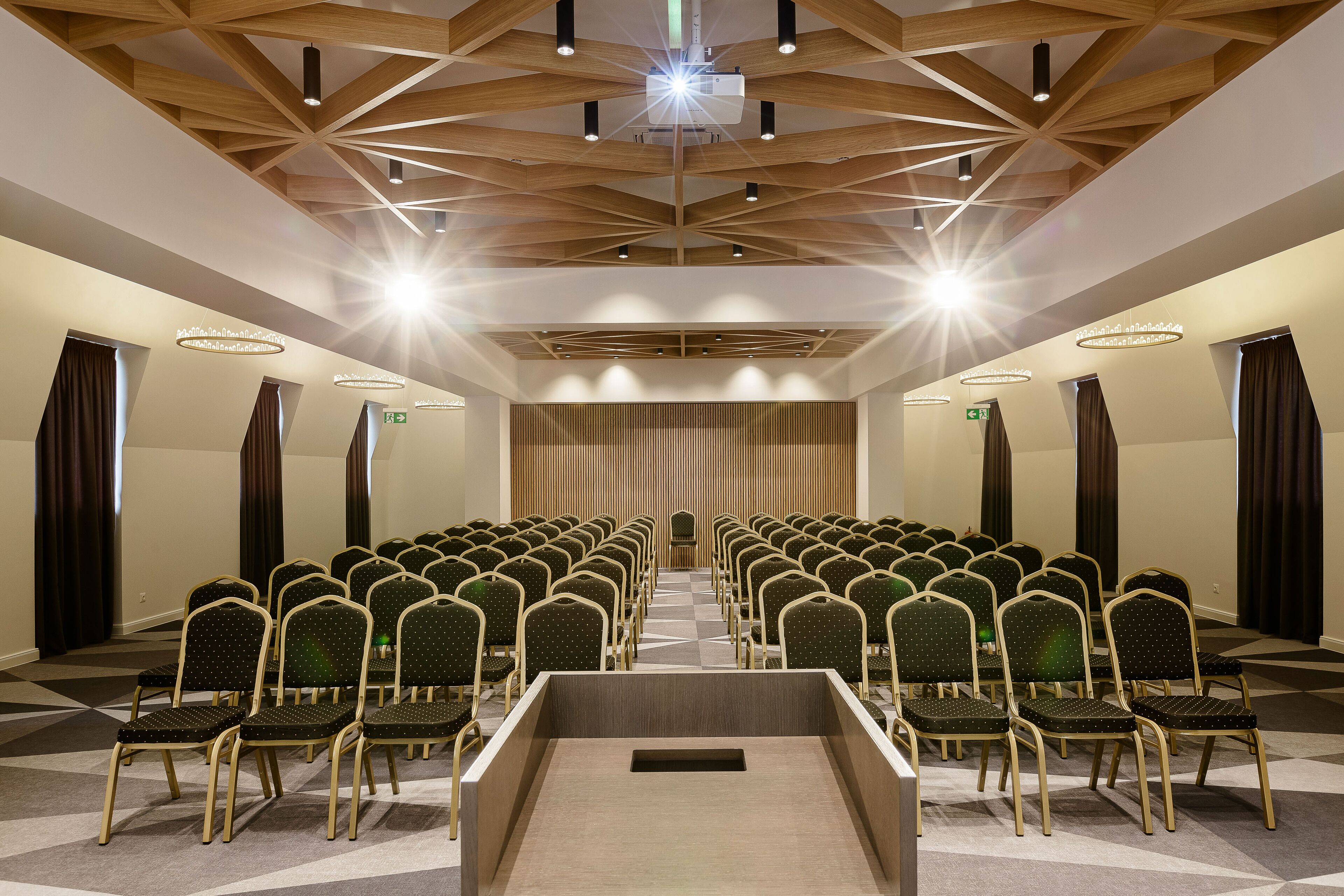 Un salón de conferencias con filas de sillas orientadas hacia un atril, vigas de madera en el techo e iluminación empotrada.