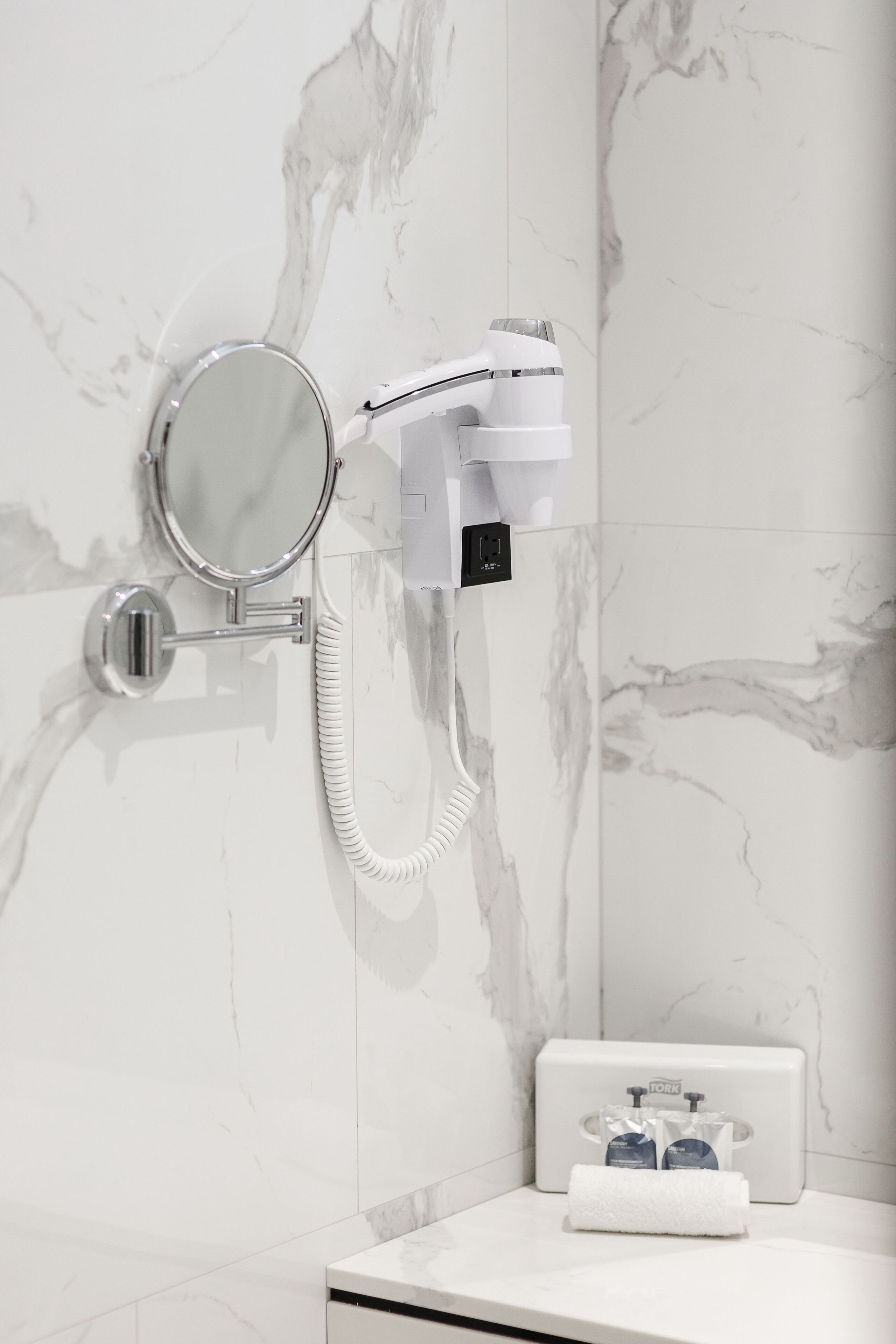 A bathroom with a marble wall, a handheld showerhead, a wall-mounted mirror, and a towel dispenser.