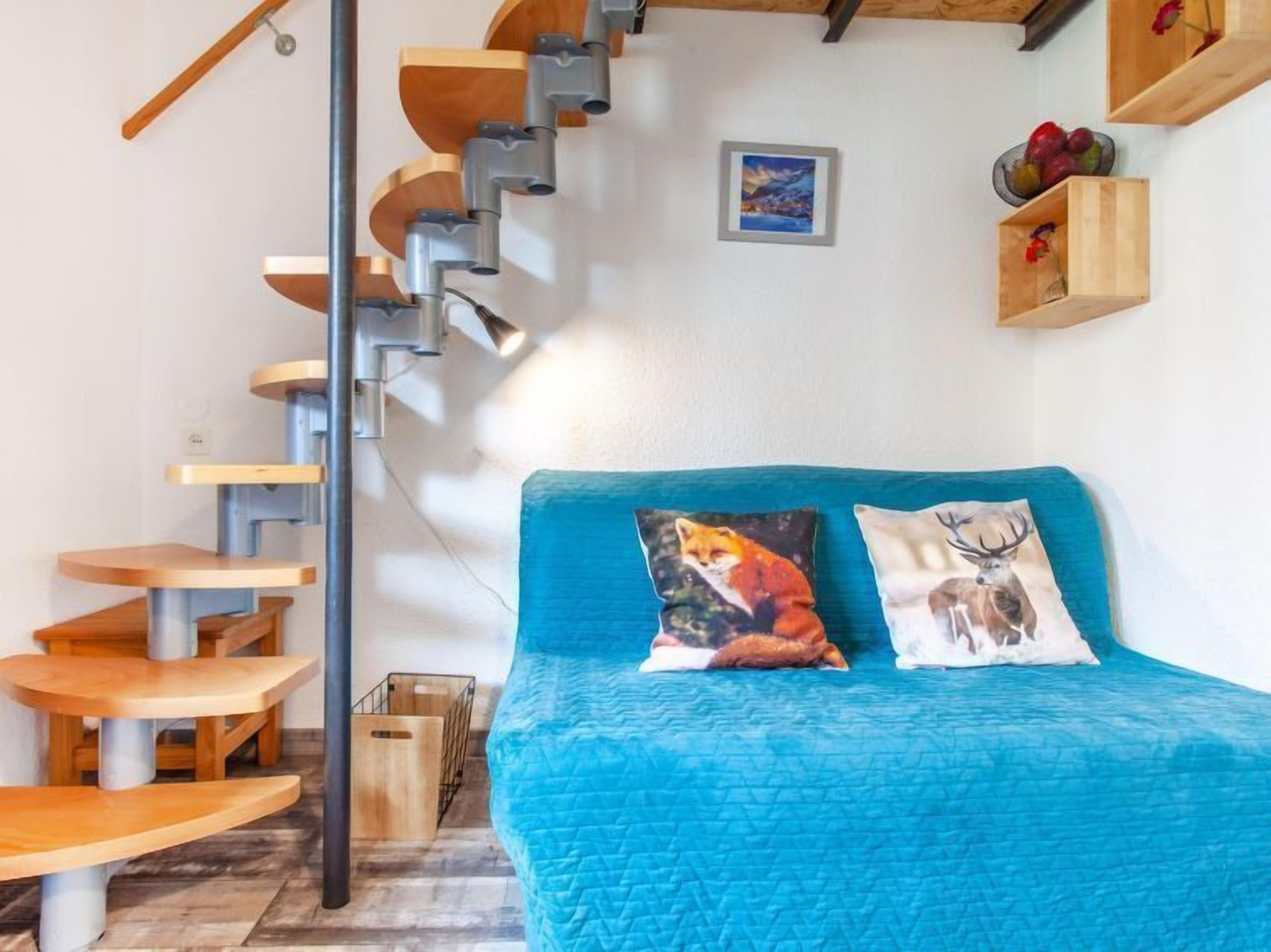 A compact room with a blue bed, a wooden staircase, and a small wooden table.