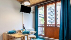 A dining area with a wooden table, blue plates, and a bottle of wine. There is a TV mounted on the wall and a view of balconies outside. - Rond Point des Pistes (Val Claret) by Interhome (Tignes)