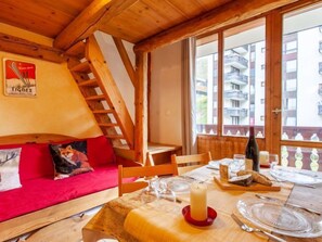 A cozy room with a wooden ceiling, a red sofa with pillows, a dining table set with plates, glasses, and a candle. A window offers a view of buildings outside. - Rond Point des Pistes (Val Claret) by Interhome (Tignes)