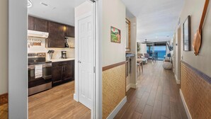 A modern kitchen with dark wood cabinets and stainless steel appliances, a hallway with wooden flooring and a view to a balcony, and a dining area with a table and chairs.