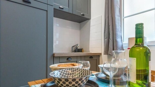 A kitchen with grey cabinets, a white tiled backsplash, a wooden table with a bowl, glasses, a bottle, and a spoon. - Le studio St Vincent by Interhome (Saint Malo)