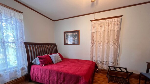 A bedroom with a wooden bed, a wooden bench, a framed picture on the wall, and lace curtains. - The Homecoming in Highmore, SD (Highmore)