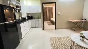 Cocina compacta con armarios blancos, refrigerador negro y un pequeño comedor con una mesa y una planta en maceta.