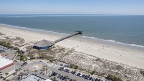 A beach with a pier extending into the ocean, a parking lot, and a building with a thatched roof. - Ms. Opal's Cottage - POOL! Pet-Friendly! Ground Level (Tybee Island)